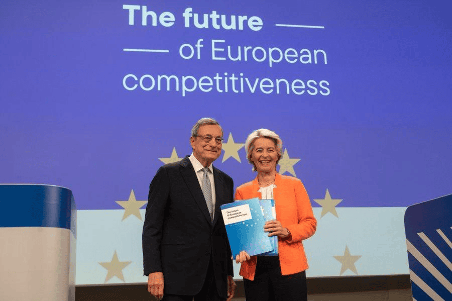 Two professionals at European competitiveness event, holding a report, with a presentation screen be.